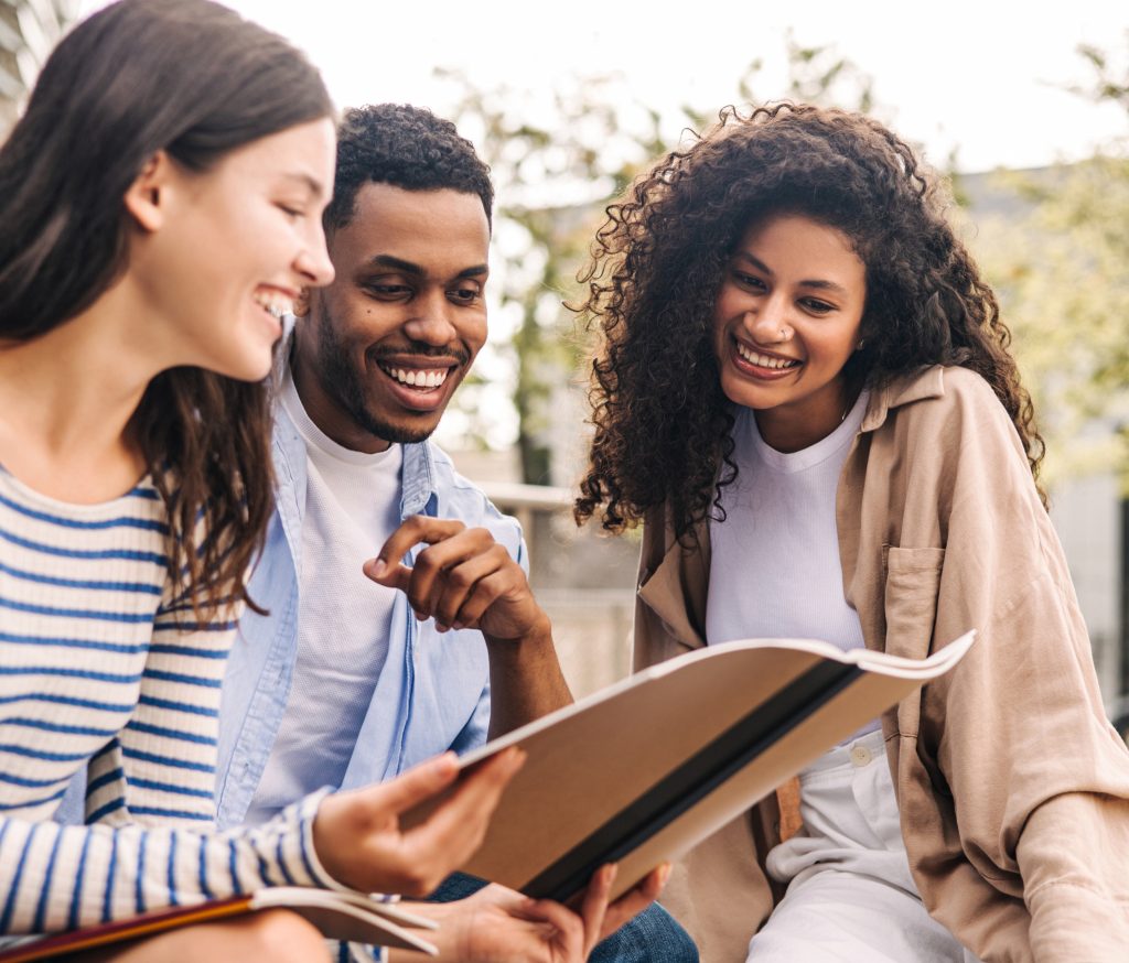 Group of young interracial students discussing homework looking at exercise book sitting on campus.